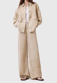 Femme portant une veste beige oversize et un pantalon large avec un haut blanc et des sandales argentées, debout sur un fond uni.