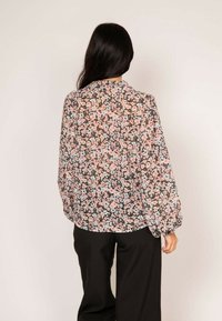 Blouse florale sur fond noir, ornée de fleurs roses et bleues, avec des manches longues et un col ; associée à un pantalon noir.