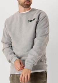 Sudadera gris hecha de tela suave con puños y dobladillo acanalados. Presenta texto en verde oscuro "GOOD" en la parte superior izquierda.