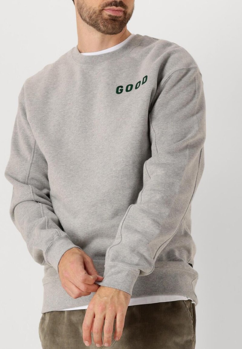 Sudadera gris hecha de tela suave con puños y dobladillo acanalados. Presenta texto en verde oscuro "GOOD" en la parte superior izquierda.
