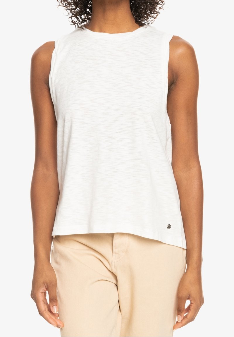 Witte mouwloze top met een textuurpatroon, ronde hals, ontspannen pasvorm en een klein logo detail rechtsonder. Gestyleerd met beige broek.