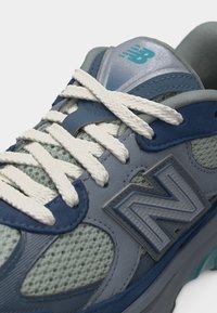 Μπλε και γκρι αθλητικό παπούτσι New Balance με λευκά κορδόνια και ύφασμα δίχτυ, με το λογότυπο στη γλώσσα και στη πλευρά, κοντινή λήψη.