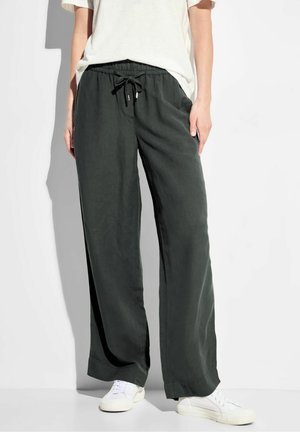 Osoba ubrana w luźne, ciemnozielone spodnie ze ściągaczem, białe trampki i białą koszulkę z krótkim rękawem, stojąca na tle jednolitego białego tła.