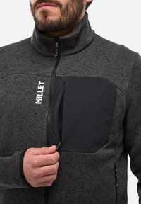 Veste en polaire gris foncé avec un col haut. Dotée d'une poche poitrine zippée noire et de deux poches latérales, elle met en avant un tissu texturé.