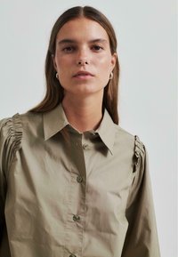 Kaki mygtukų marškiniai su struktūrizuotu apykakle, susirinkimo detalėmis ant pečių siūlių ir lygiomis, šiek tiek blizgiomis audinio tekstūromis.