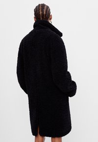 Manteau noir en fausse laine d'agneau avec un col montant, des manches larges, une fente latérale à l'ourlet, présentant une texture moelleuse et un design surdimensionné.