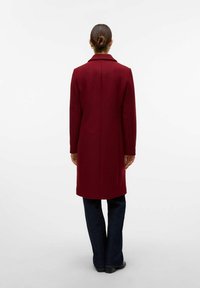 Lungo cappotto bordeaux con taglio dritto, colletto a revers e texture liscia. Indossato sopra pantaloni in denim scuri, caratterizzati da un design minimalista.