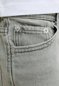 Κοντινό πλάνο στην τσέπη γκρι τζιν με μεταλλικά καρφιά και λευκή ραφή πουκάμισου από πάνω.