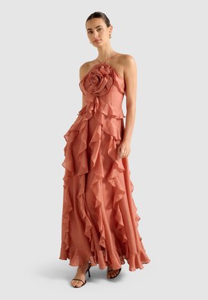 Kvinde iført en ærmeløs, støvet rose maxi-kjole med flæser og en stor blomsterdetalje på brystet, parret med sorte strop-sandaler.