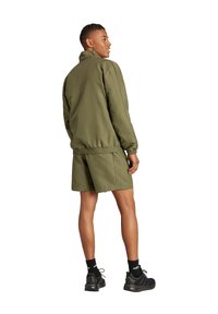 Olivgrüne Sportjacke mit elastischen Bündchen und Saum, kombiniert mit passenden Shorts. Getragen mit schwarzen Sneakers und Socken. Rückansicht.