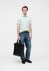 Ανοιχτό μπλε πόλο πουκάμισο, μπλε τζιν, μαύρη δερμάτινη τσάντα tote με κάθετες ραφές και μαύρα παπούτσια. Το μοντέλο στέκεται με το πρόσωπο προς τα εμπρός μπροστά σε ουδέτερο φόντο.