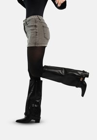 Persoon die grijze denim shorts draagt, zwarte panty's en zwarte leren laarzen tot aan de knie met een lage hak, en een been naar achteren optilt.