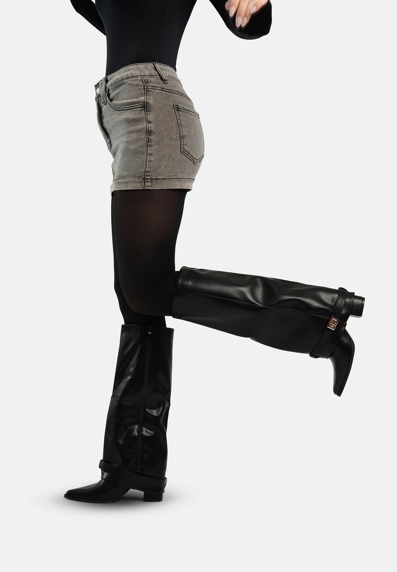 Persoon die grijze denim shorts draagt, zwarte panty's en zwarte leren laarzen tot aan de knie met een lage hak, en een been naar achteren optilt.