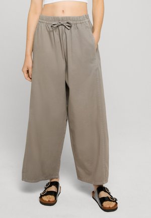 Osoba oblečená v loose sivých šnúrkových nohaviciach a čiernych sandáloch s prackou, stojaca proti jednoliatemu svetlosivému pozadiu.