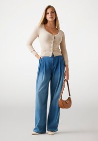 Beige ribbed cardigan with buttons, paired with high-waisted blue wide-leg jeans. Model holds a tan shoulder bag and wears pointed shoes.