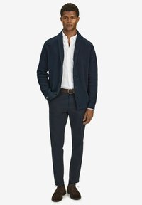 Marineblå stickad kofta med sjalkrage över en vit skjorta med knappar, kombinerad med mörka byxor och bruna snörskor, mot en neutral bakgrund.