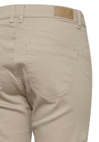 Pantalons en coton beige montrant des poches arrière avec un design incurvé, des détails cousus et une étiquette en cuir marron texturé sur la taille.