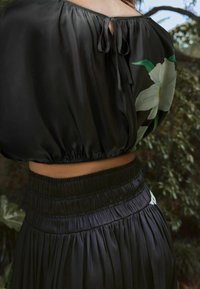 Haut court en satin noir avec un col froncé, un détail de nœud dans le dos et un imprimé floral. Taille élastique sur une jupe plissée assortie.