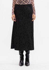 Saia maxi preta com um padrão texturizado, apresentando um acabamento em tecido suave. Combinada com botas pretas brilhantes até ao joelho.
