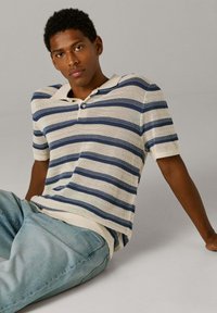 Jeune homme assis par terre portant un polo en maille à manches courtes rayé blanc et marine et un jean bleu clair, sur un fond uni.