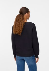 Sudadera negra con un corte relajado, cuello redondo y mangas largas, combinada con jeans azules. El material parece suave con una textura lisa.