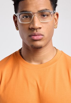 Junger Mann mit klaren rechteckigen Brillen, goldener Halskette, goldenen Creolen und orangefarbenem T-Shirt mit Rundhalsausschnitt, neutraler Gesichtsausdruck.