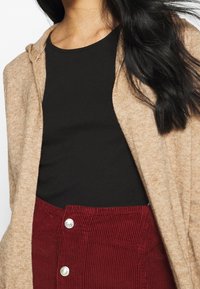 Béžový pletený kardigan s kapucí přes černý top, doplněný vínovou manšestrovou sukní se stříbrnými knoflíky a svislými žebrováními.