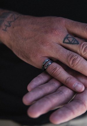 Rokas ar tetovējumiem, vienai uz vidējā pirksta ir melns un sudraba teksturēts gredzens tumšā fonā.