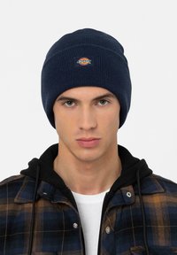 Joven con un gorro de punto azul oscuro con el logo de Dickies, sudadera negra y chaqueta a cuadros azul y marrón sobre una camiseta blanca.