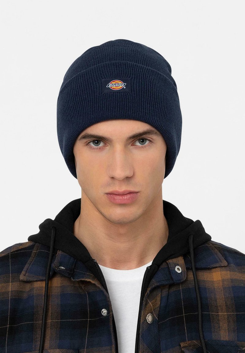 Joven con un gorro de punto azul oscuro con el logo de Dickies, sudadera negra y chaqueta a cuadros azul y marrón sobre una camiseta blanca.
