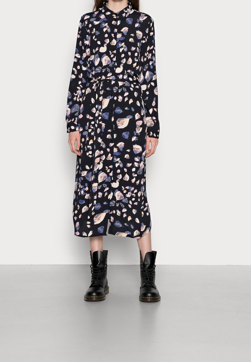Femme portant une robe midi noire à manches longues avec des motifs de feuilles roses et bleues, taille ceinturée, et des bottines noires lacées de style militaire.