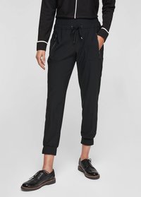 Zwarte joggers met een trekkoord in de taille, zijzakken met rits en taps toelopende boorden. Draagt shiny zwarte schoenen. Gladde stoftextuur.