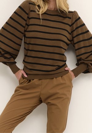 Person iført en brun sweater med sorte, vandrette striber og puffede ærmer, matchet med brune bukser, hænderne i lommerne.