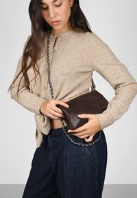 Clutch de cuero texturizado marrón con una cadena plateada. Llevado con un suéter beige y jeans oscuros, mostrando un diseño compacto.