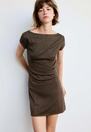 Jeune femme portant une robe marron à carreaux ajustée à manches courtes, debout devant un fond clair uni.