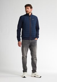 Gilet imbottito blu navy con frontale trapuntato, maniche in maglia, chiusura a zip completa e un'etichetta. Indossato con jeans grigi e scarpe da ginnastica bianche.
