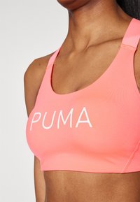 Rožinė sportinė liemenėlė, pagaminta iš lygaus audinio, su plačiu dirželiu, storomis petnešomis ir ryškiu baltu "PUMA" logotipu priekyje.
