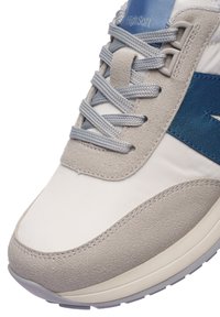 Gros plan sur une basket beige et blanche avec un logo et des lacets bleus, présentant un mélange de matériaux en suède et en matériaux lisses, avec une étiquette "High Soft" sur la languette.