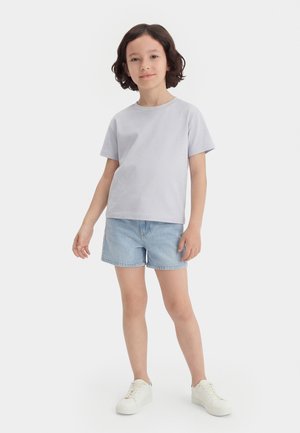 Rövid sötét hajú gyermek áll, világosszürke pólót, világoskék farmer rövidnadrágot és fehér tornacipőt visel, fehér háttér előtt.