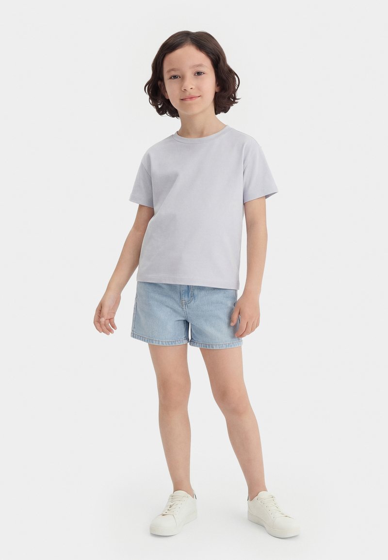 Παιδί με κοντά σκούρα μαλλιά όρθιο, που φοράει ανοιχτό γκρι μπλουζάκι, ανοιχτά μπλε τζιν σορτσάκι και λευκά αθλητικά παπούτσια σε λευκό φόντο.