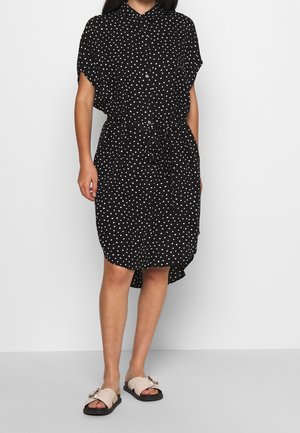 Mujer con vestido negro hasta la rodilla con lunares blancos y sandalias beige de tipo slide con suelas negras.
