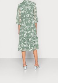 Vestido midi floral de tejido transparente verde, con falda en capas, mangas tres cuartos y un cinturón de lazo en la cintura. Combinado con tacones de color azul claro.