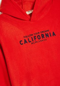 Sudadera roja con capucha, bolsillo frontal, cordones en la capucha y texto bordado que dice "SIGUE TUS SUEÑOS CALIFORNIA MCMLXVIII". Textura suave.