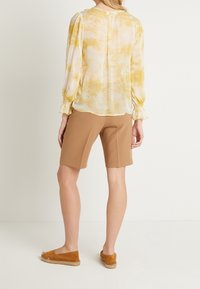 Gul tie-dye blus med samlade ärmar, i kombination med bruna shorts och bruna espadrillosandaler. Minimalistisk bakvy med slät textur.