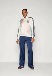 Balta zip-up striukė su pastelinių geometrinių raštų, juodomis juostelėmis; tamsiai mėlyni kelnių su šoninėmis juostelėmis; sportiniai batai. Sportinės aprangos komplektas.