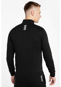 Felpa nera con colletto alto, maniche lunghe e dettaglio a striscia bianca sul retro. Realizzata in un materiale liscio e elasticizzato.