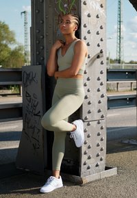 Conjunto de athleisure verde-claro com um top cropped e leggings de cintura alta, combinado com ténis brancos, frente a um fundo de coluna metálica.