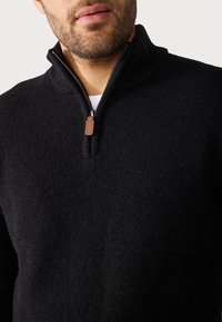 Jersey de punto negro con cuello de media cremallera, tejido texturizado y tirador de cremallera de cuero marrón. Presenta un ajuste ceñido y un diseño casual.