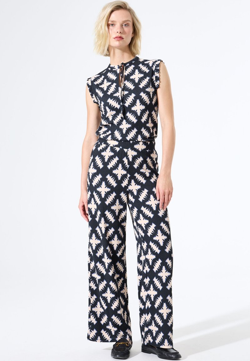 Maritieme jumpsuit met een roomkleurig geometrisch patroon, voorzien van korte ruches mouwen, een keyhole-detail aan de voorkant en wijde broekspijpen. Gladde stoftextuur.