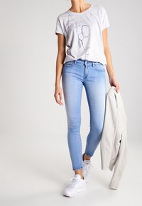  camiseta de color gris claro con texto gráfico en azul marino, combinada con unos jeans ajustados de color azul claro. El modelo lleva zapatillas blancas y sostiene una chaqueta ligera.